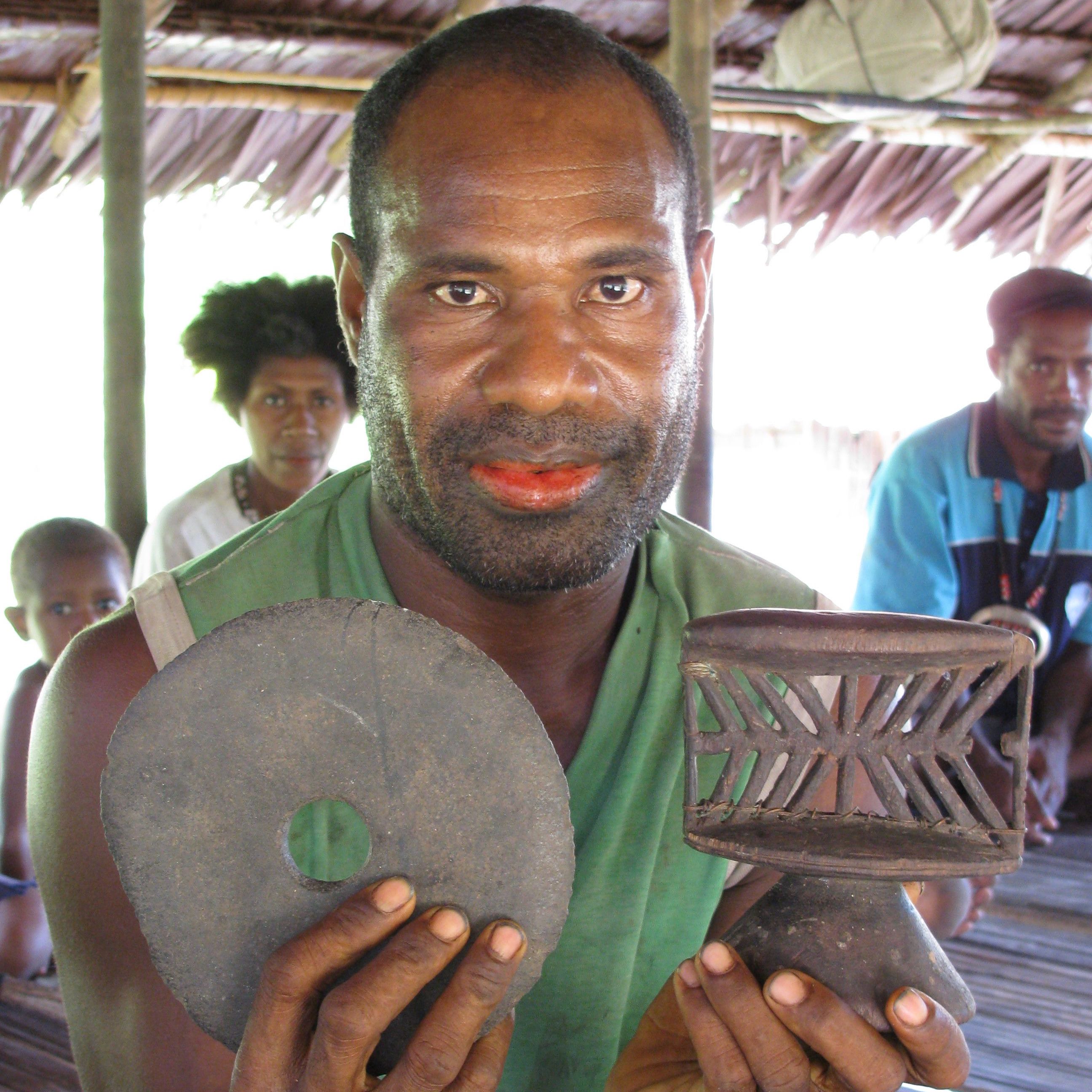 Nice stone disc club head and old neckrest—notice wire repair—Collingwood Bay, Oro Province, Papua New Guinea.  Equally visually interesting is the green from the shirt contrasting with the red betel-stained lips.
